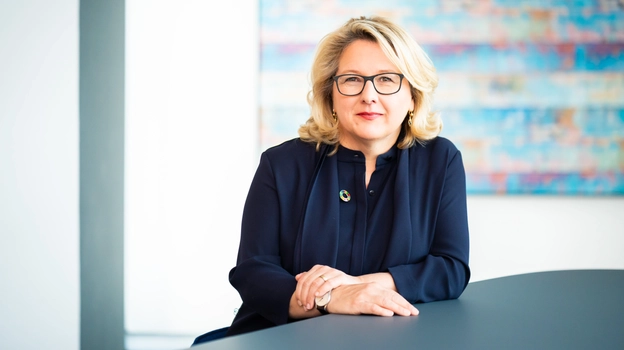 	Federal Minister for Economic Cooperation and Development Svenja Schulze at a dark grey desk in front of a white wall with a colourful painting on it