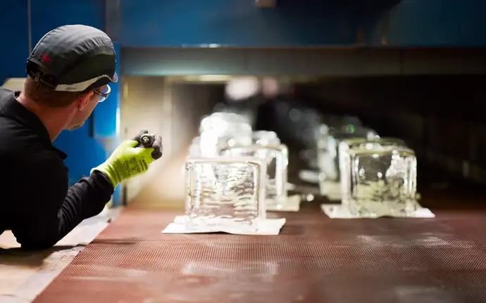 A man with a protective cap shines a torch on numerous glass cubes from a machine on a conveyor belt