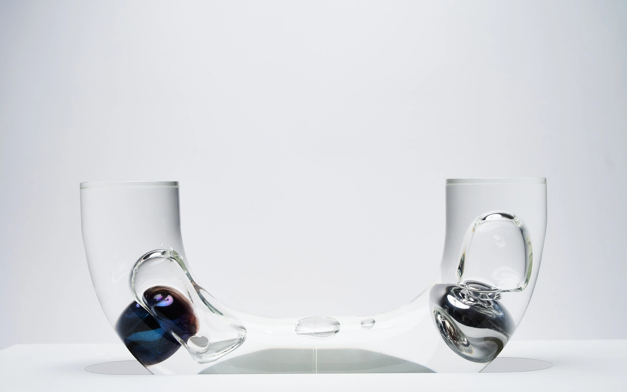 Clear U-shaped glass vessel with dark blue and black bulbous ends against a plain white background