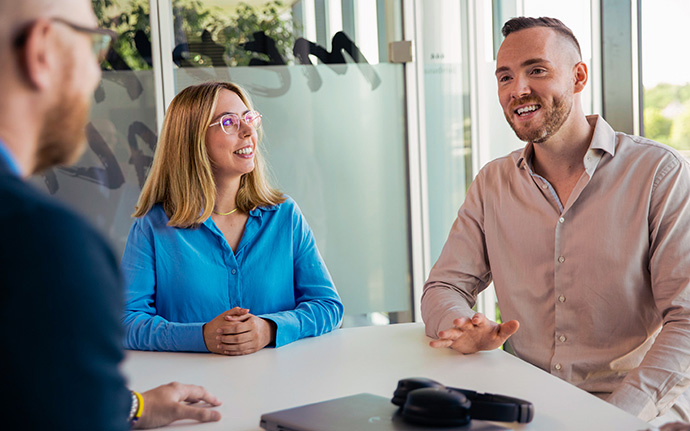 Zwei Personen sitzen an einem Tisch und unterhalten sich, während ein weiterer Gesprächspartner im Vordergrund steht.