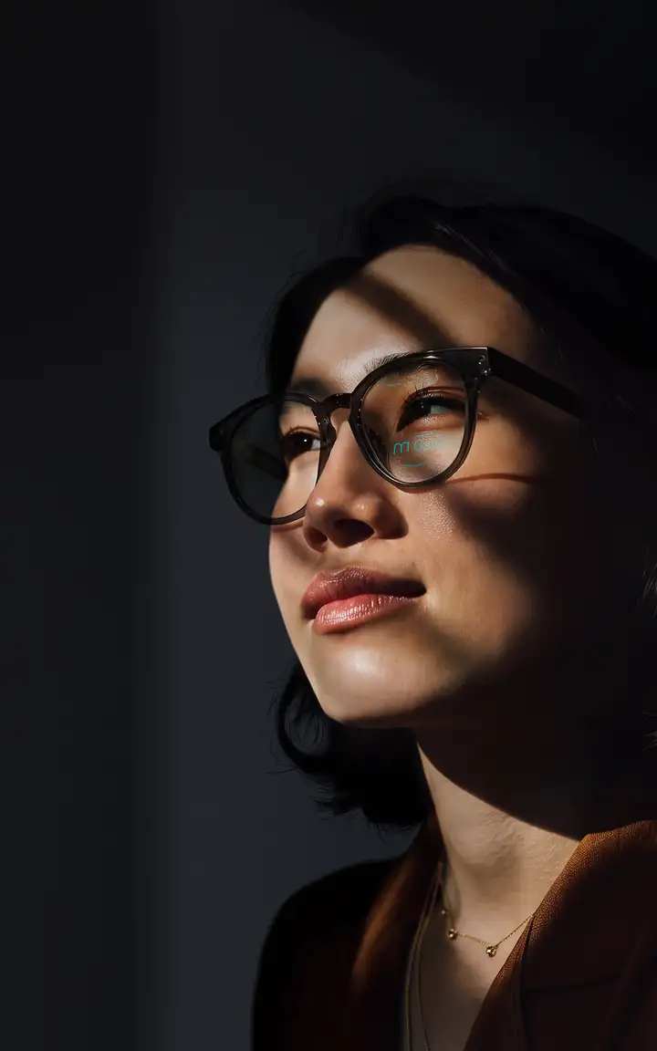 A close-up of a person wearing glasses, lit dramatically with shadow patterns across the face, against a dark background.