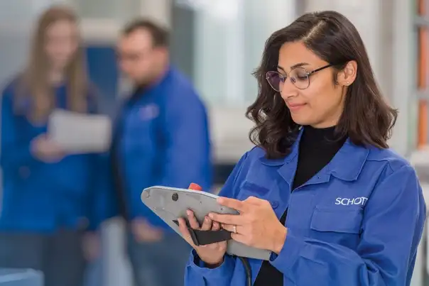 Eine Person hält ein Tablet in den Händen und arbeitet daran, während im Hintergrund zwei weitere Mitarbeitende in blauer Arbeitskleidung miteinander sprechen.