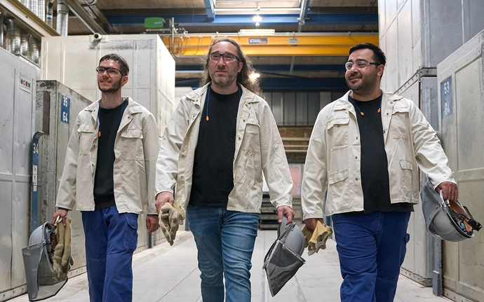 Drei Personen in heller Schutzkleidung gehen nebeneinander durch eine industrielle Umgebung und tragen Helme und Handschuhe bei sich.