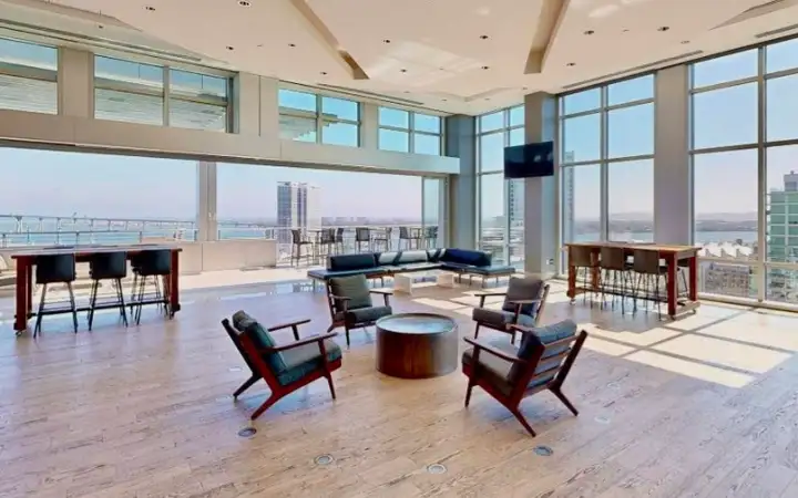 A modern lounge area with large windows overlooking a cityscape and harbor, featuring seating and tables.