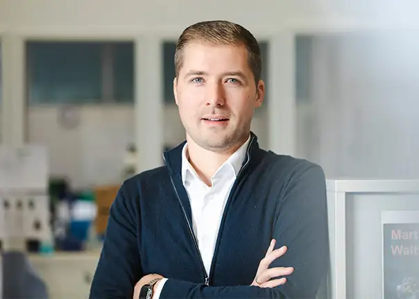 Eine Person in Business‑Kleidung steht mit verschränkten Armen in einem Büro.