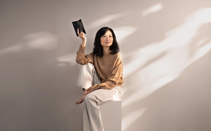 Eine Person sitzt auf einem Podest in einem hellen Studio und hält ein Handy in der Hand.