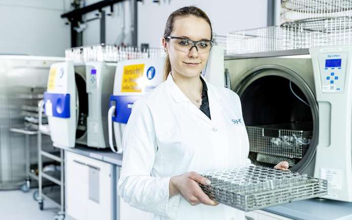 Frau im Labor mit Schutzbrille und weißem Arbeitskittel hält ein Nest aus Pharmafläschchen in der Hand.