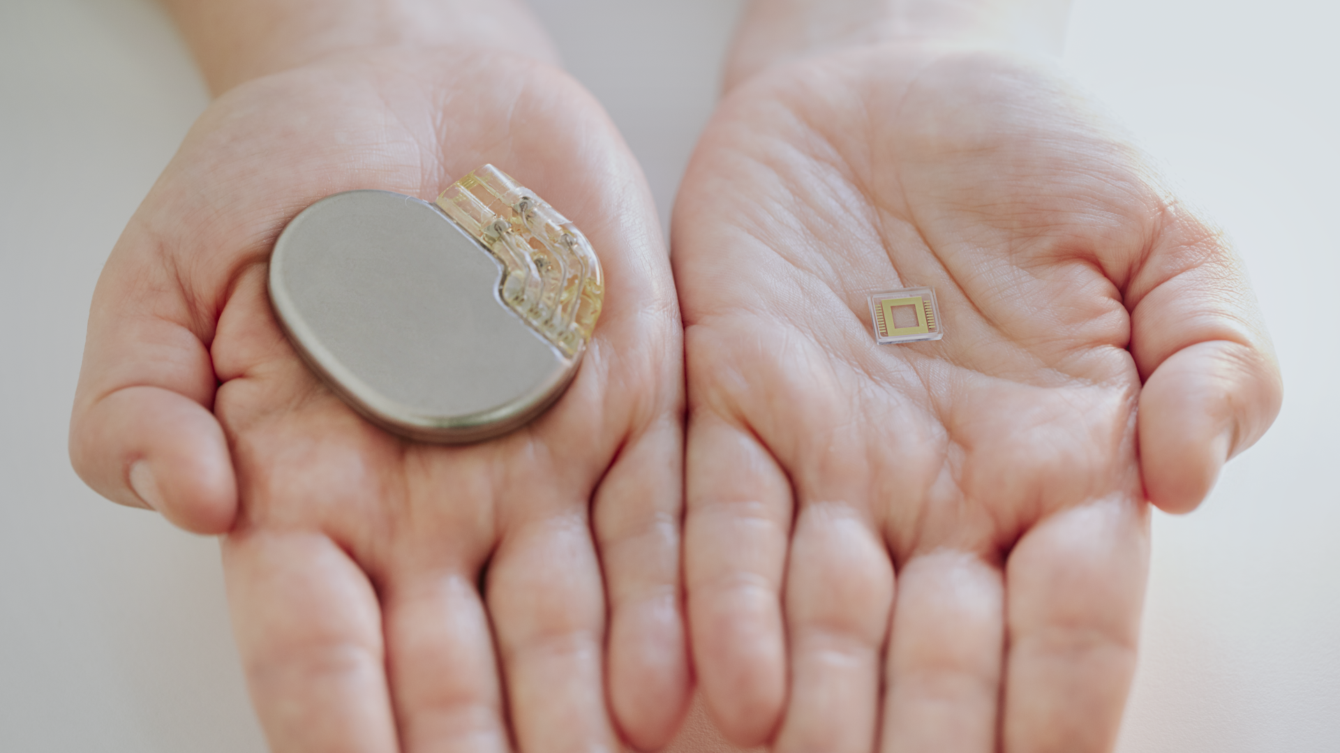 A person holding a large implantable medical device and a small implant featuring Primoceler Proteon ™ glass encapsulation, for size comparison.