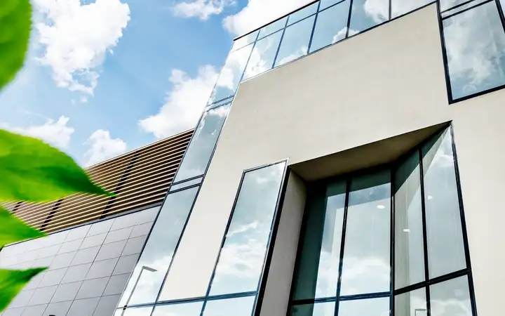 Modern building with large glass windows and clean architectural lines under a bright blue sky with scattered clouds, with a green leaf in the foreground.