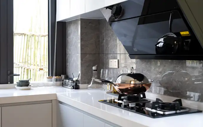 Modern kitchen with a black extractor hood made of glass, sleek white countertops, a gas glass hobtop, and various kitchen utensils near a large window.