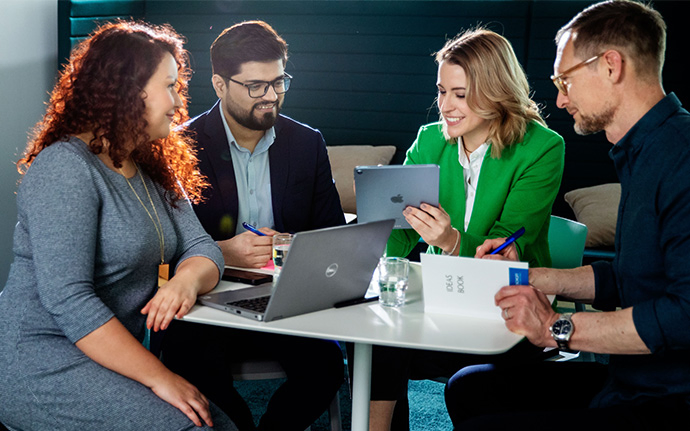 Vier Personen sitzen in einer kleinen Gruppe an einem Tisch und arbeiten gemeinsam mit Laptop, Notizbuch und Tablet.