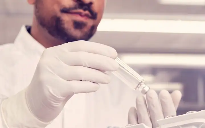 Two lab technicians in cleanroom suits inspect a cartridge tray of small glass vials in a laboratory.