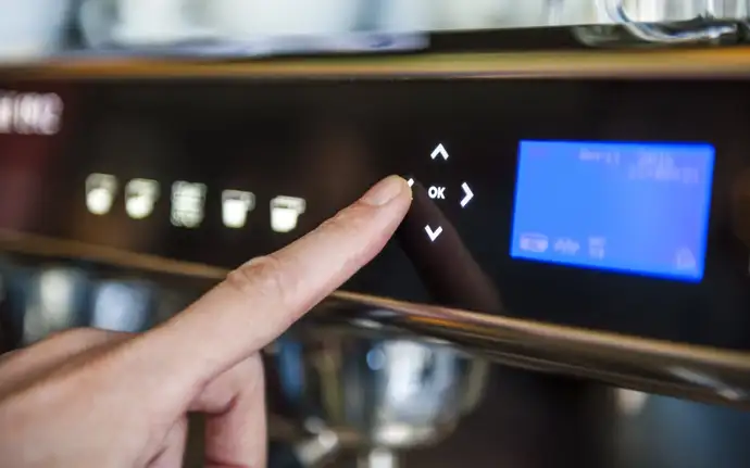 Close-up of a coffee machine glass control panel with a finger pressing the “OK” button, surrounded by navigation arrows and an illuminated blue digital display.