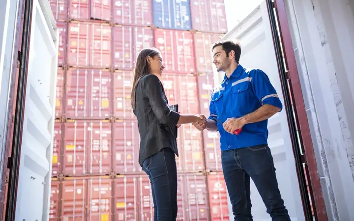 Woman in gray suit shakes hand of man next to large stack of containers