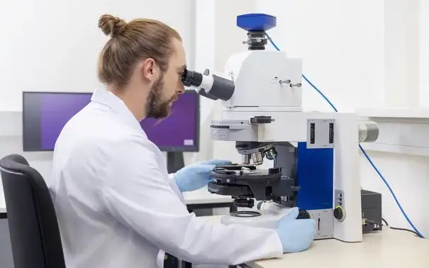 A scientist wearing a lab coat and gloves is using a microscope, focusing on a sample in a laboratory setting.