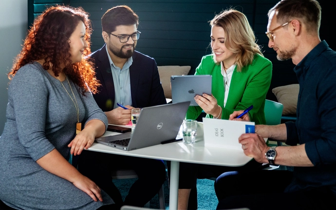 Vier Personen sitzen gemeinsam an einem Tisch und arbeiten mit Laptop, Tablet und Notizmaterialien an einer Aufgabe.