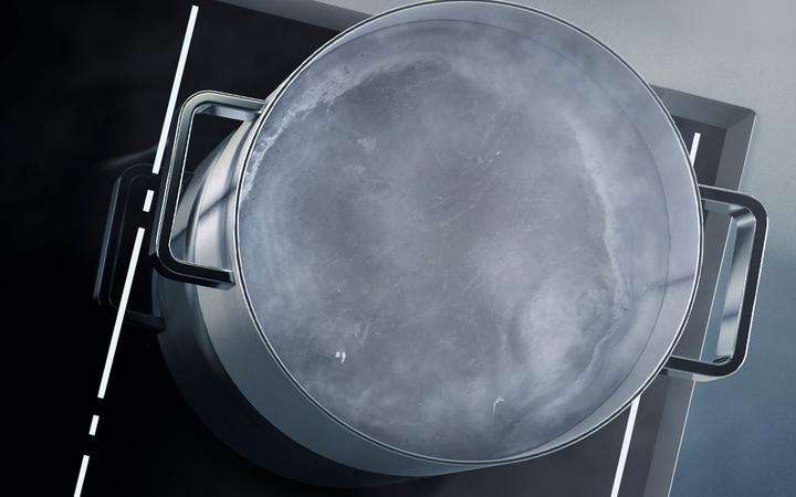 Ecke einer SCHOTT CERAN® Kochfläche von oben mit weiß leuchtenden Lichtstreifen und Topf mit kochendem Wasser darauf.