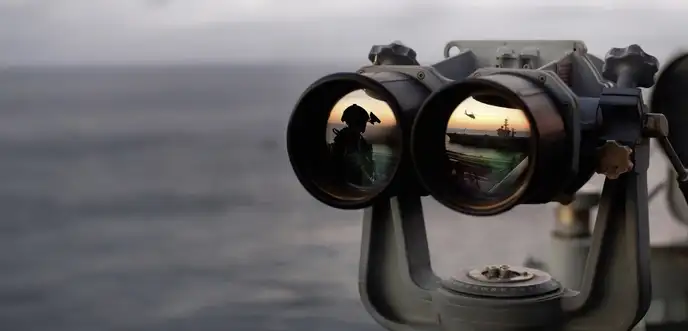 Binoculars reflecting a soldier and a helicopter over an aircraft carrier at sunset
