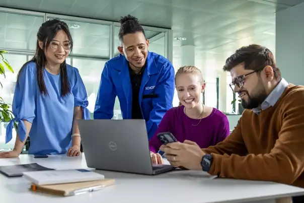 Vier Personen arbeiten gemeinsam an einem Tisch und betrachten Informationen auf einem Laptop.