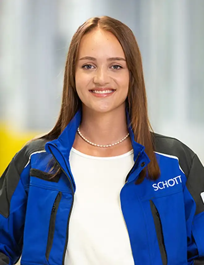 Eine Person trägt eine blaue SCHOTT‑Arbeitsjacke und steht in einer hellen Produktionshalle.