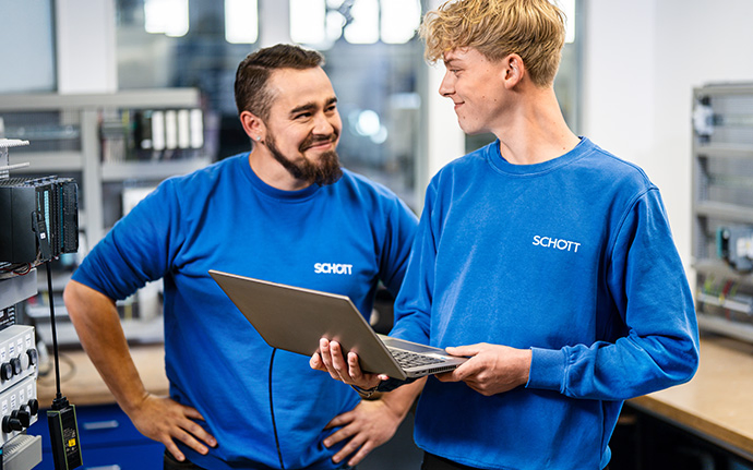 Zwei Mitarbeitende in blauen Arbeitsoberteilen stehen in einer Werkstatt und besprechen etwas an einem Laptop.
