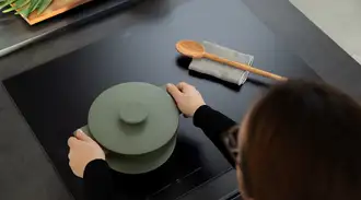 Person placing a green pot on a black matte SCHOTT CERAN® induction cooktop, with a wooden spoon on a cloth beside it.