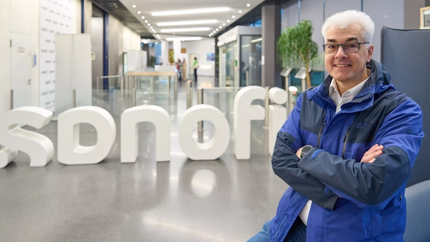 A man standing with arms crossed, smiling, in front of a large "Sanofi" logo inside a modern office building