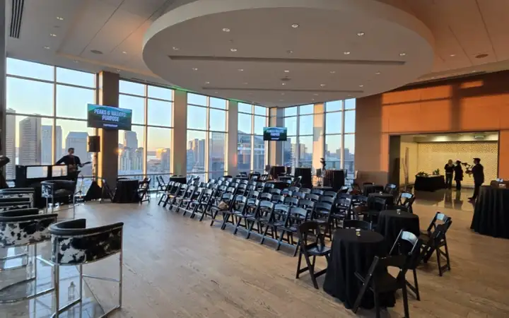 A large event space with black chairs arranged in rows and round tables, featuring a wall of windows overlooking a downtown skyline.