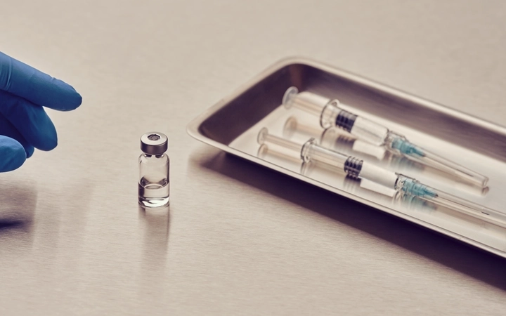Gloved hand reaching toward a small vial beside three syringes on a metal tray
