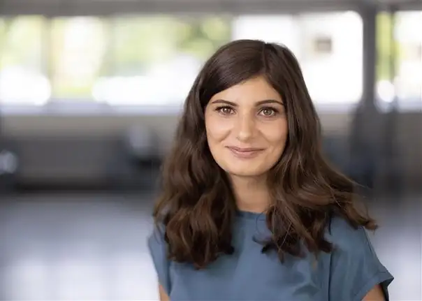 Das Bild zeigt eine Person mit schulterlangem, welligem Haar, die in einem großen, hellen Innenraum mit weichen Lichtreflexionen und unscharfem Hintergrund steht.