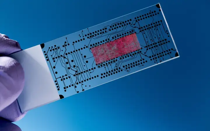 Gloved hand holding a glass lab-on-a-chip on a blue background