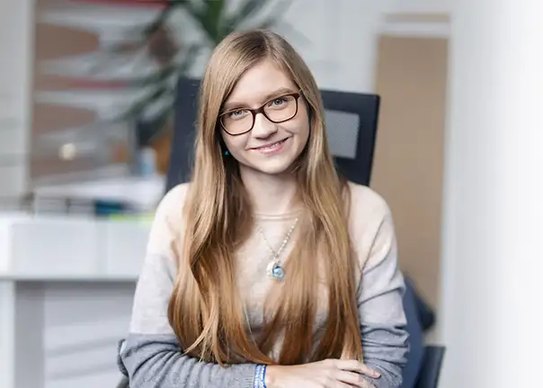 Das Bild zeigt eine Person, die an einem Schreibtisch sitzt und in einem Bürobereich fotografiert wurde, wobei lange hellbraune Haare, ein zweifarbiges Oberteil und ein auffälliger Anhänger sowie Armbänder sichtbar sind.