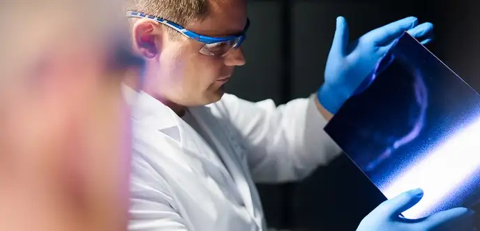 Eine Person in Labor Kleidung und Handschuhen hält eine glänzende Platte prüfend gegen eine Lichtquelle.