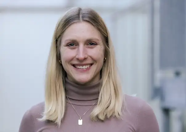 Das Bild zeigt eine Person, die im Freien vor einem hellen, unscharfen Hintergrund steht und ein langärmliges, eng anliegendes Oberteil sowie eine dunkle Hose mit einem sichtbaren Gürtel trägt, ergänzt durch eine Halsketten.
