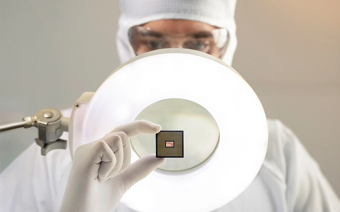 Eine Person in Schutzanzug und Handschuhen hält ein kleines elektronisches Bauteil vor eine starke Lichtquelle.