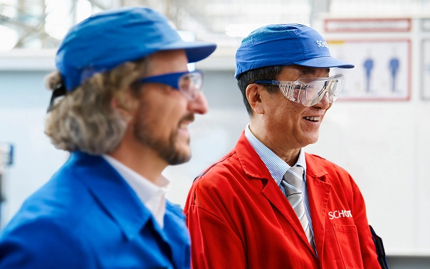 Zwei Personen in farbigen Arbeitsjacken und Schutzkappen stehen nebeneinander in einer Produktionshalle.