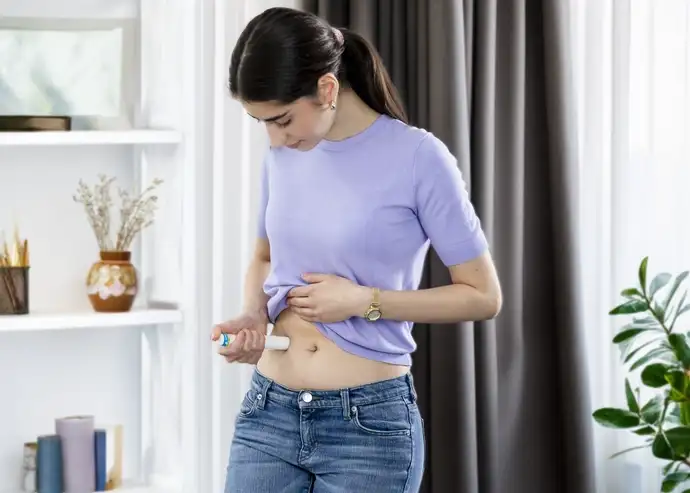 A young woman in jeans and a shirt is standing in the living room, self-injecting medication via an autoinjector in her right hand.