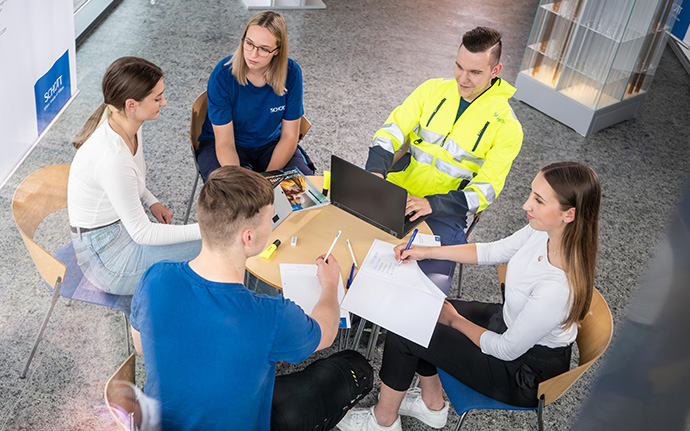 Fünf junge Menschen sitzen um einen Tisch und arbeiten gemeinsam an Dokumenten und einem Laptop.