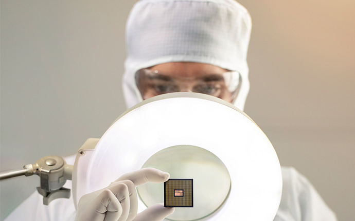 Eine Person in Schutzkleidung hält einen kleinen Mikrochip vor eine helle Lupenleuchte, sodass die feinen Strukturen sichtbar werden.