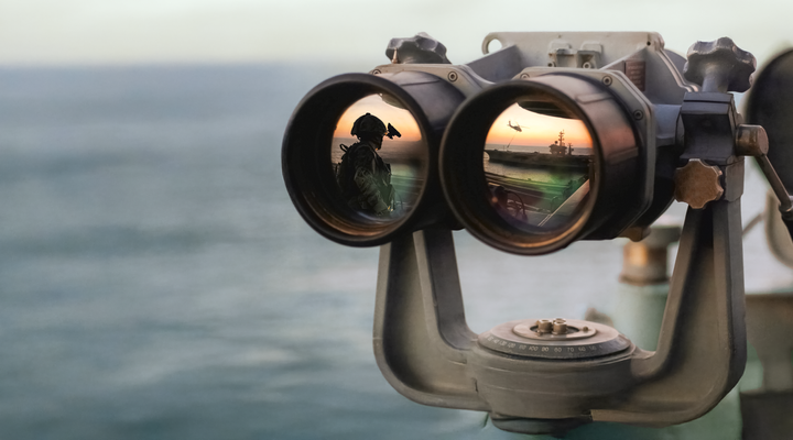 Binoculars reflecting a soldier and a helicopter over an aircraft carrier at sunset
