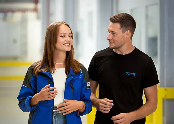 Zwei Personen gehen durch einen hellen Arbeitsbereich und unterhalten sich, wobei beide Firmenkleidung tragen.