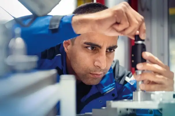 Eine Person arbeitet präzise an einer Maschine und justiert mit einem Werkzeug ein technisches Bauteil in einer industriellen Umgebung.