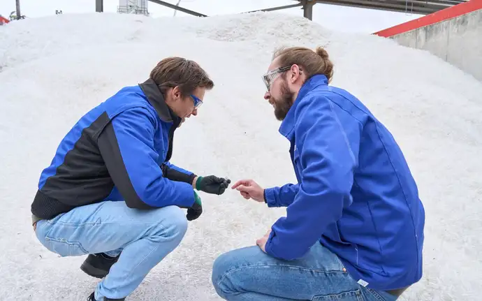 Zwei Männer in blauer SCHOTT Arbeitsschutzkleidung mit Sicherheitsbrille knien vor dem SCHOTT Glasfläschchen Scherbenlager.