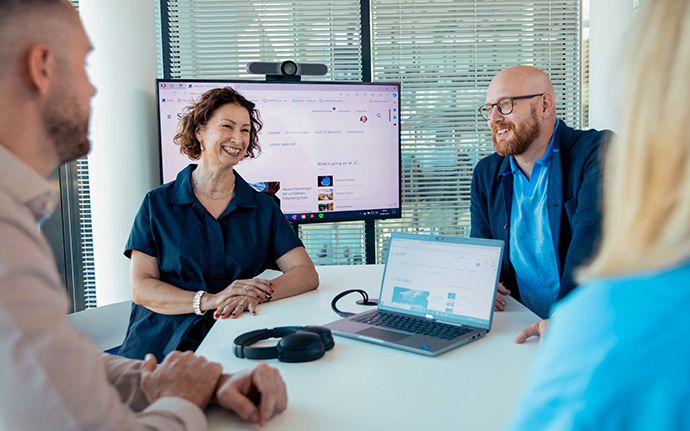 Vier Personen sitzen an einem Tisch vor einem großen Monitor und sprechen über die Inhalte, die auf einem Laptop und dem Bildschirm angezeigt werden.