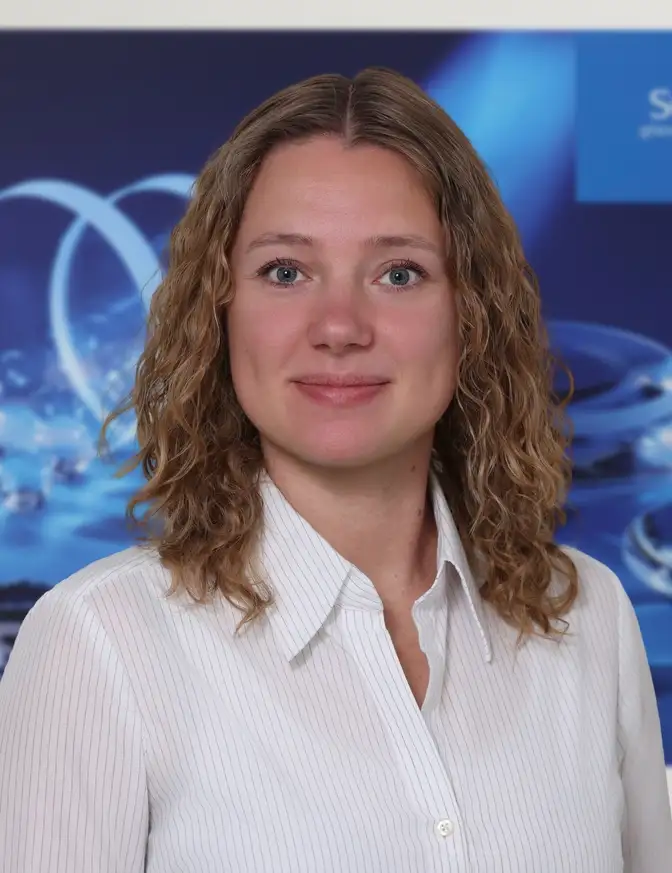 Eine Person mit lockigem, schulterlangem Haar steht vor einem blauen Firmenposter der Marke SCHOTT, auf dem Glasobjekte abgebildet sind.