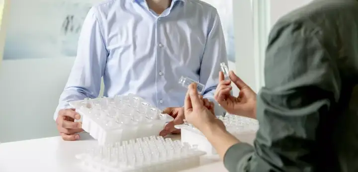Two people handling small transparent laboratory vials over trays filled with similar sample tubes on a work table, suggesting sample sorting or preparation in a lab or clinical setting