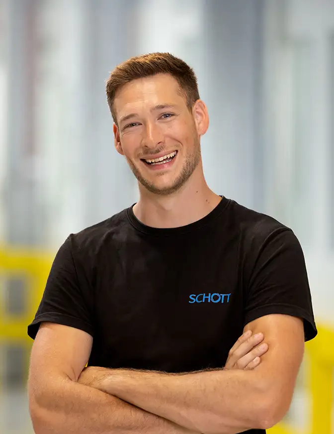 Eine Person trägt ein schwarzes SCHOTT‑T-Shirt und steht mit verschränkten Armen in einer hellen Produktionsumgebung.