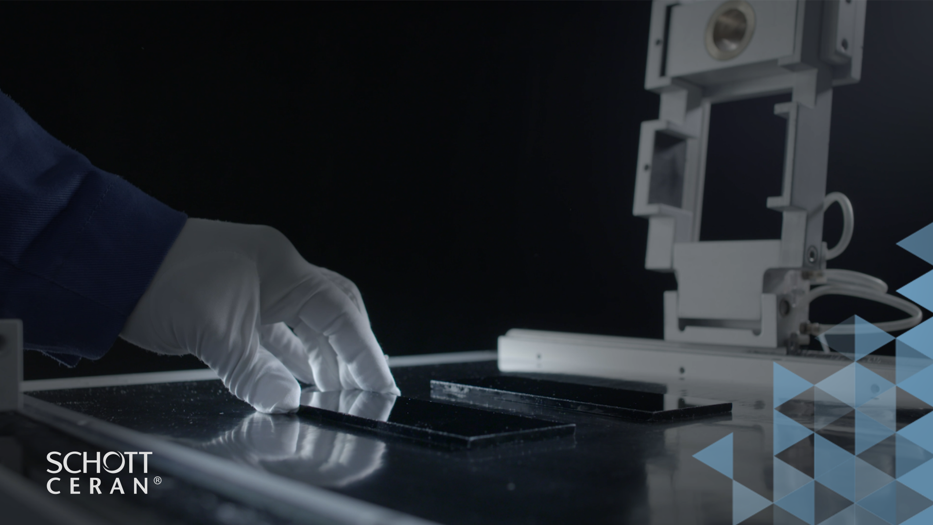 A gloved hand touches a glass-like material on a testing table. A piece of equipment is visible in the background.