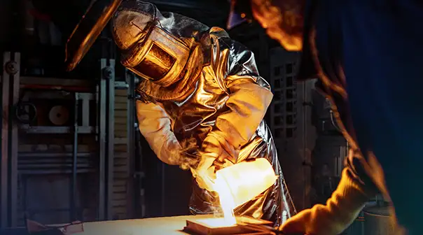 Eine Person in Schutzkleidung gießt in einer Werkstatt flüssiges Material in eine Form.