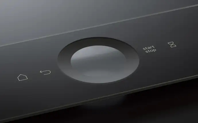 Close-up of a sleek black glass control panel featuring a circular touch dial and minimalist icons for home, back, start/stop, and timer.